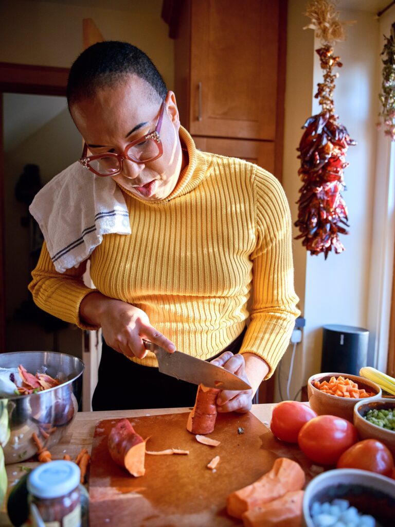 Black woman with yellow turtleneck and pink glasses chops sweet potato with dish towel over her shoulder.
