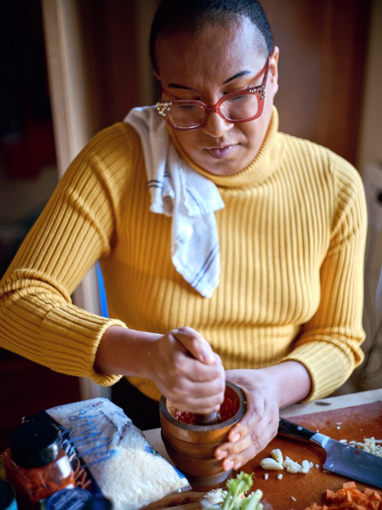 Black woman with yellow turtleneck and pink glasses grinds ingredients in mortar and pestle, with dish towel over her shoulder.