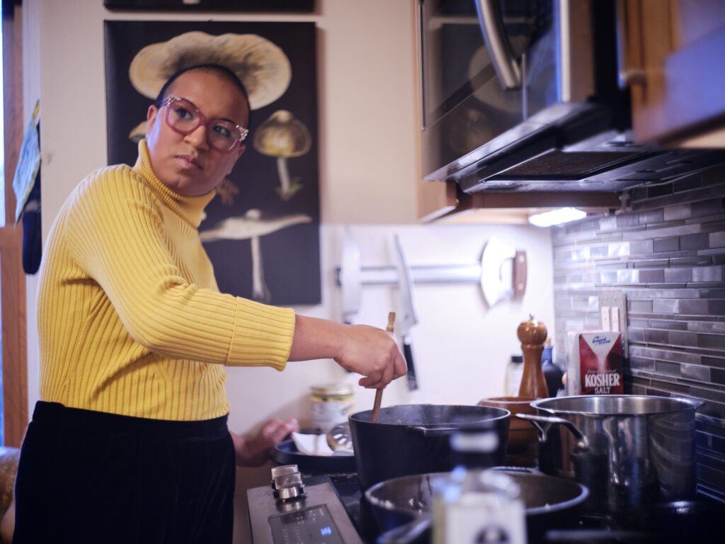 Black woman with yellow turtleneck and pink glasses stirs pot on stove while looking over shoulder.