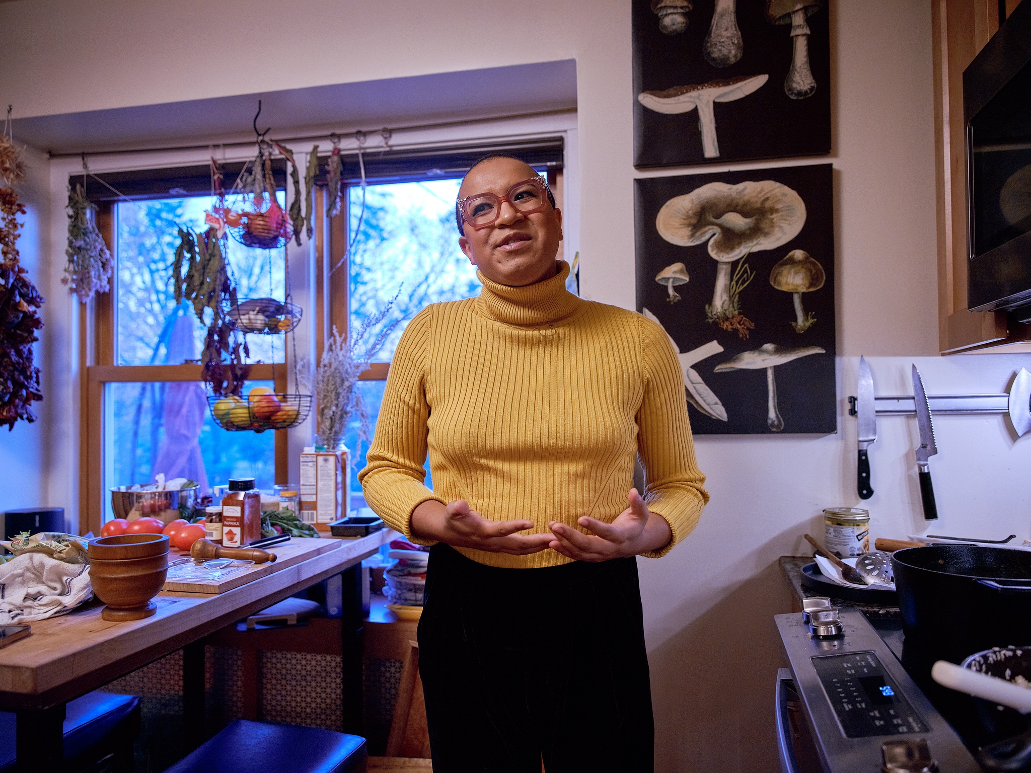 Black woman with yellow turtleneck and pink glasses stands between stove and kitchen island, gesturing with open hands.