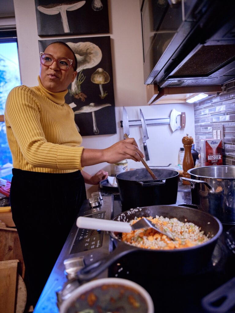 Black woman with yellow turtleneck and pink glasses stirs pot on the stove, looking over her shoulder.