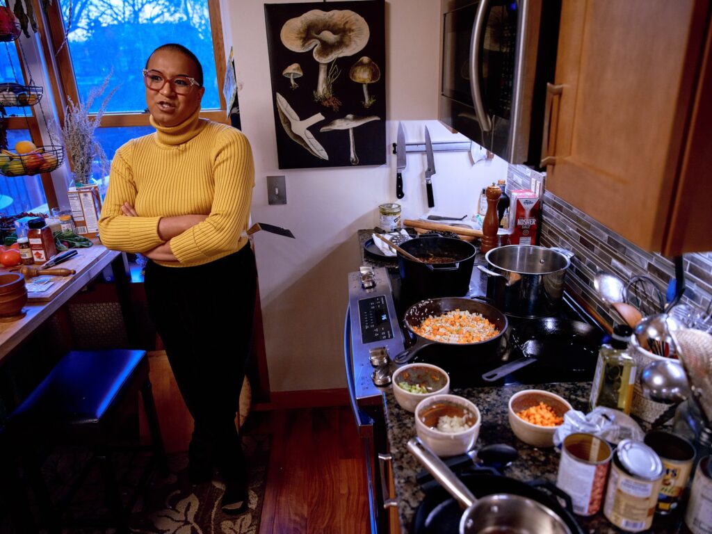 Black woman with yellow turtleneck and pink glasses stands next to stove with arms folded.
