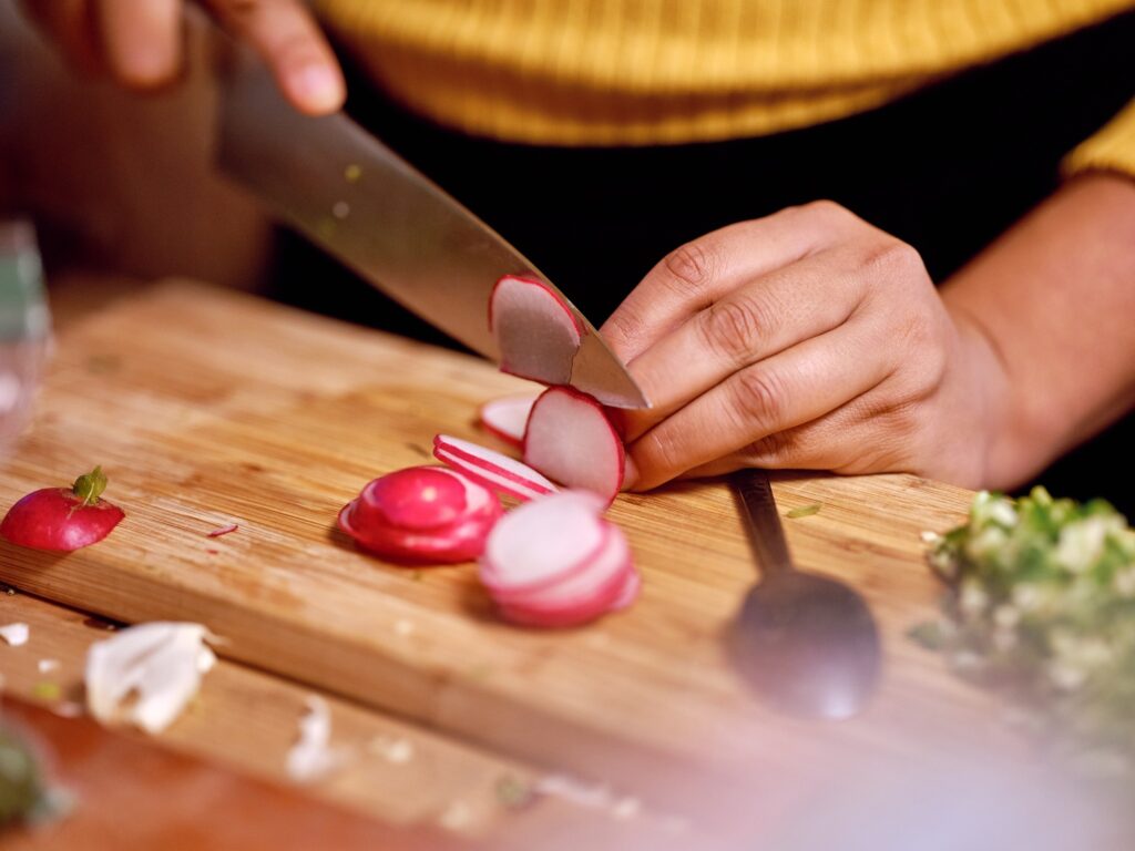 Hands thinly slice radish on wooden cutting board.