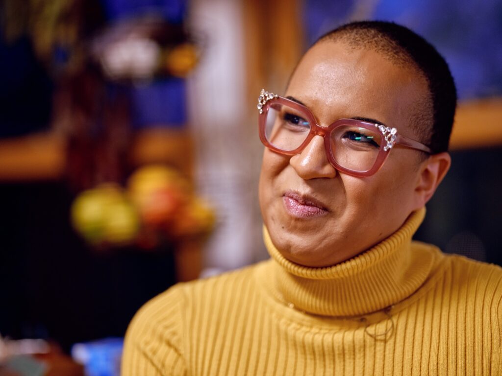 Black woman with yellow turtleneck and pink glasses smiles with closed lips.