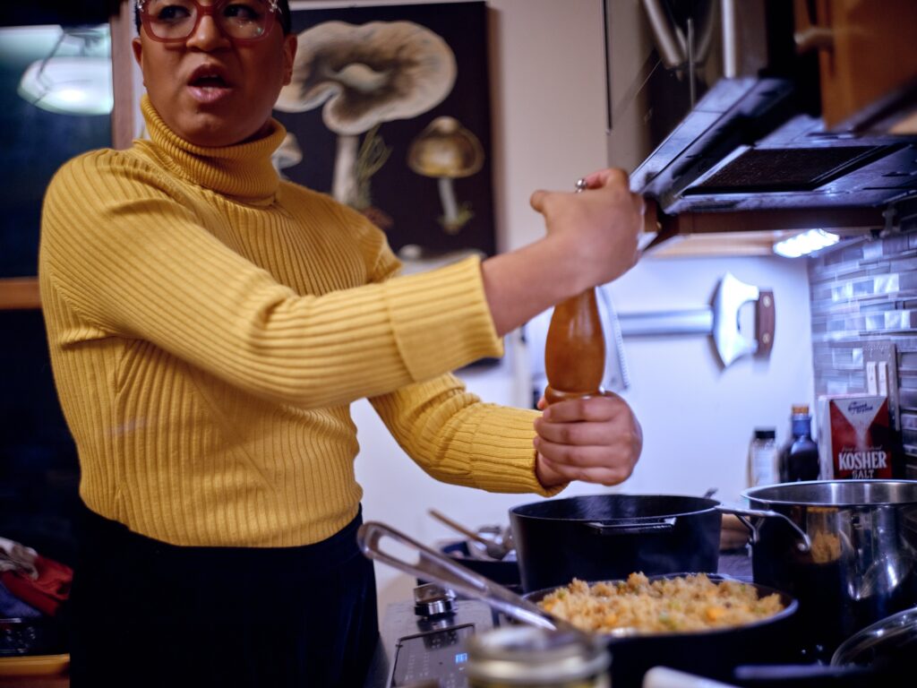 Black woman with yellow turtleneck and pink glasses grinds pepper into dish on stove.