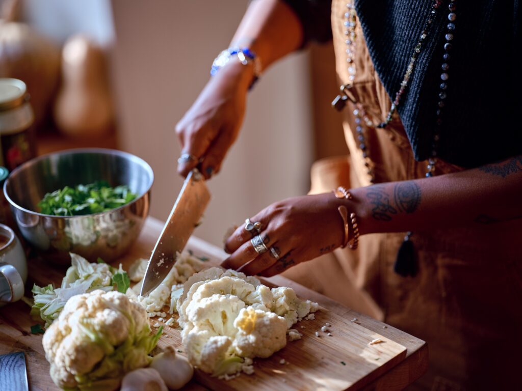 Hands with dark skin and jewelry chop cauliflower on wooden cutting board.