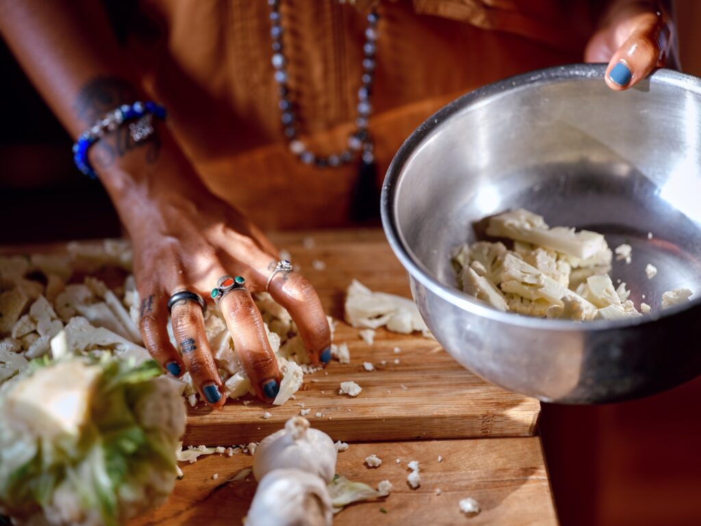 Hands with dark skin and jewelry move chopped cauliflower from cutting board to metal bowl.