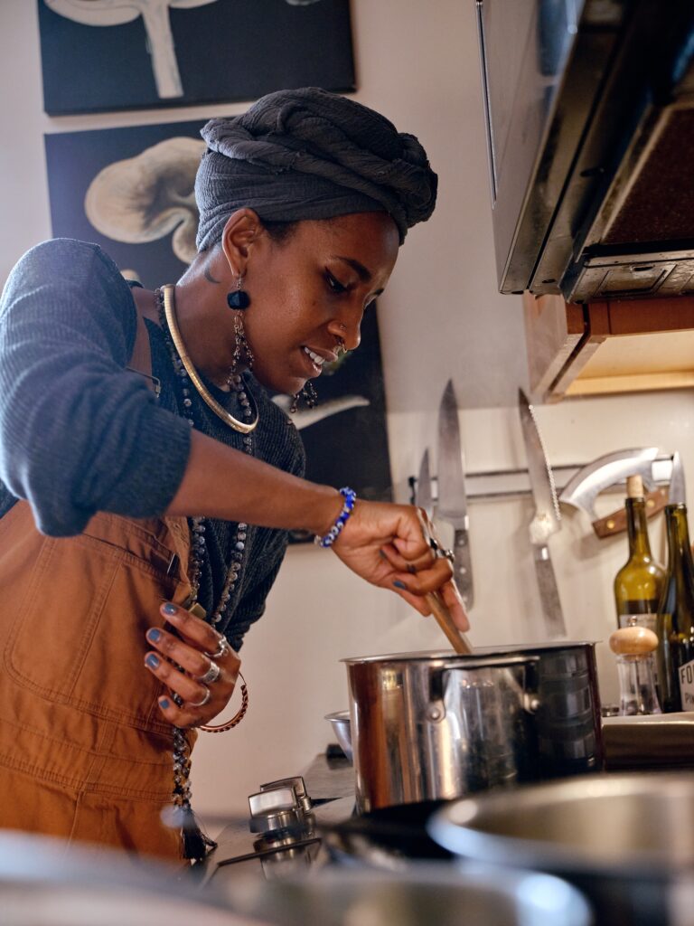 Black woman wearing overalls, jewelry, and gray headwrap stirs pot on stove.