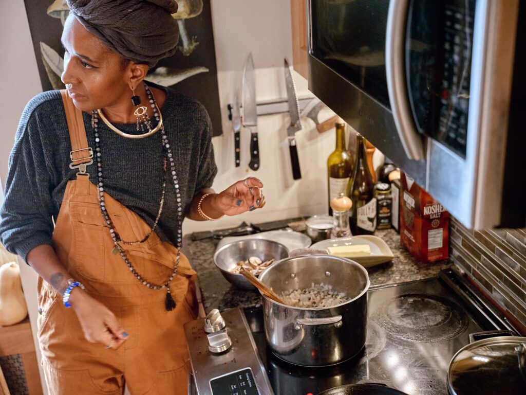 Black woman wearing overalls, jewelry, and gray headwrap stands over pot on stove and looks back over her shoulder.