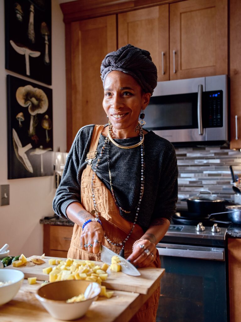 Black woman wearing overalls, jewelry, and gray headwrap smiles while chopping potatoes.