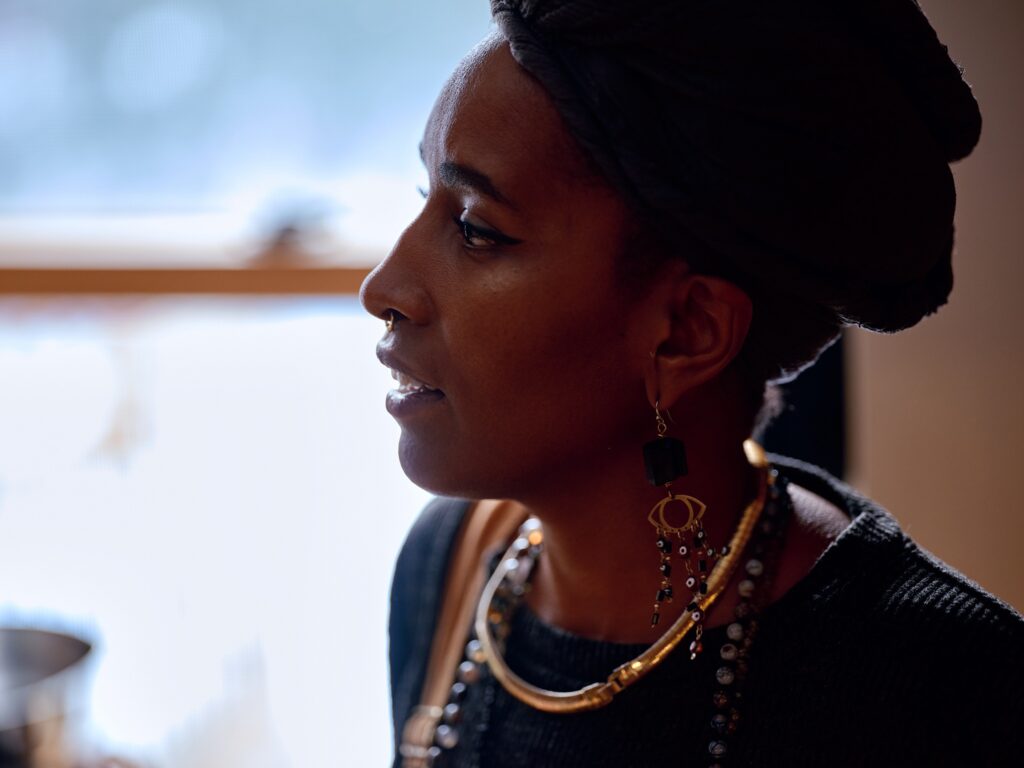 Black woman wearing overalls, jewelry, and gray headwrap in profile and slight silhouette.