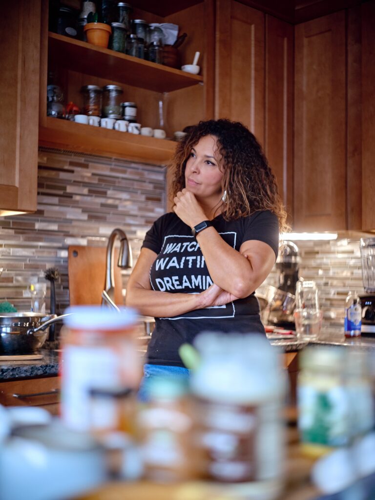 Person with medium skin and dark curly hair stands in kitchen and rests her chin on her hand.