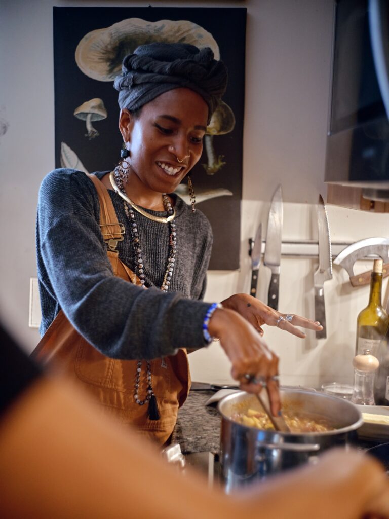 Black woman wearing overalls, jewelry, and gray headwrap smiles and stirs pot on stove.