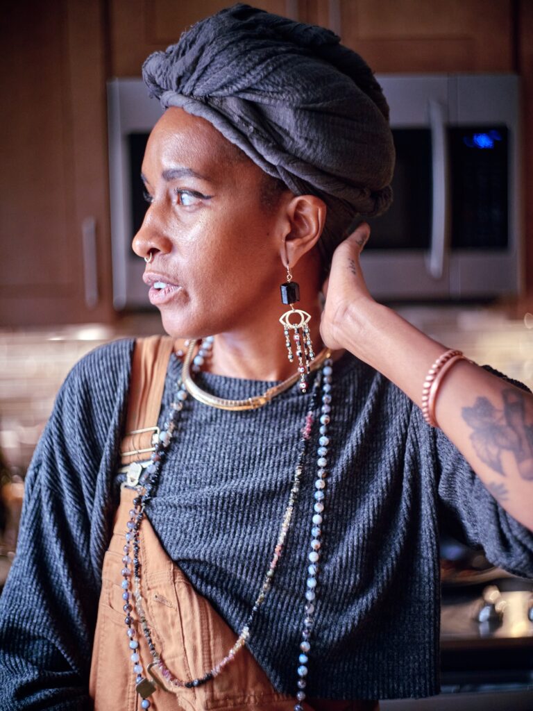 Black woman wearing overalls, jewelry, and gray headwrap touches the back of her neck and looks over her right shoulder.
