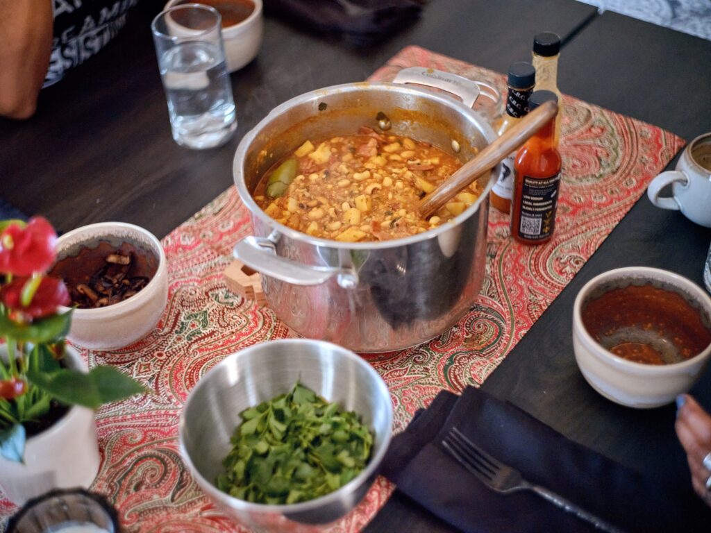 Table set with large pot of food and wooden spoon, bottles of hot sauce, bowl of cilantro, red paisley table cloth, and table settings.