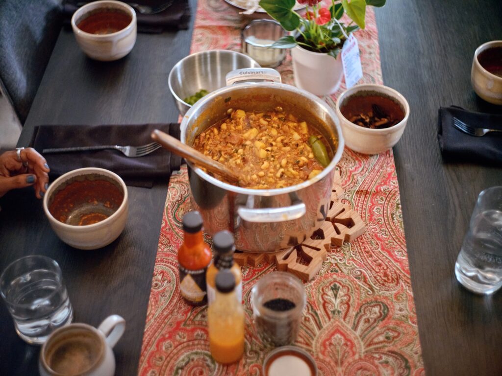 Table set with large pot of food and wooden spoon, bottles of hot sauce, bowl of cilantro, red paisley table cloth, and table settings.