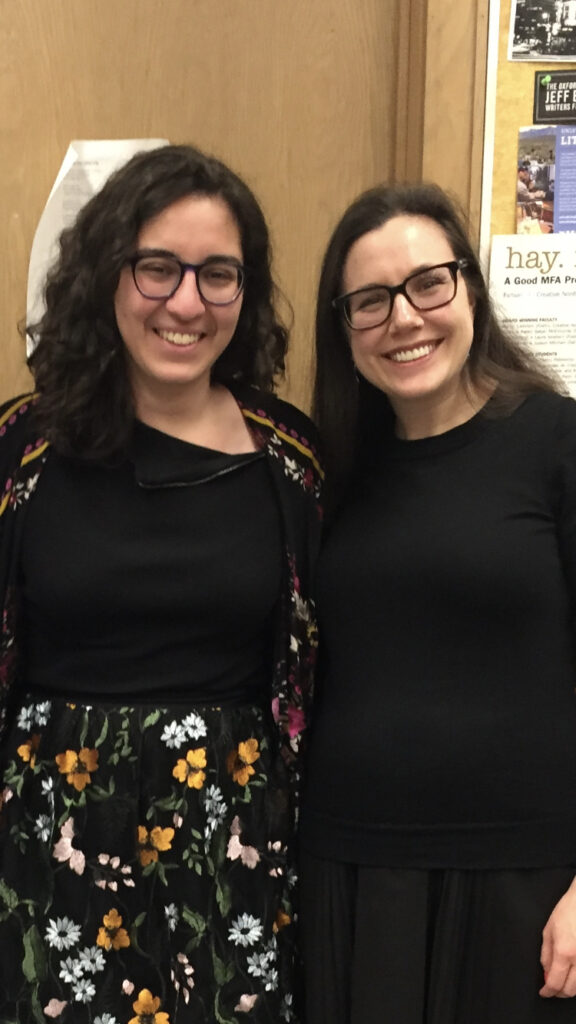 Two people with glasses and long dark hair lean close to each other and smile at the camera.