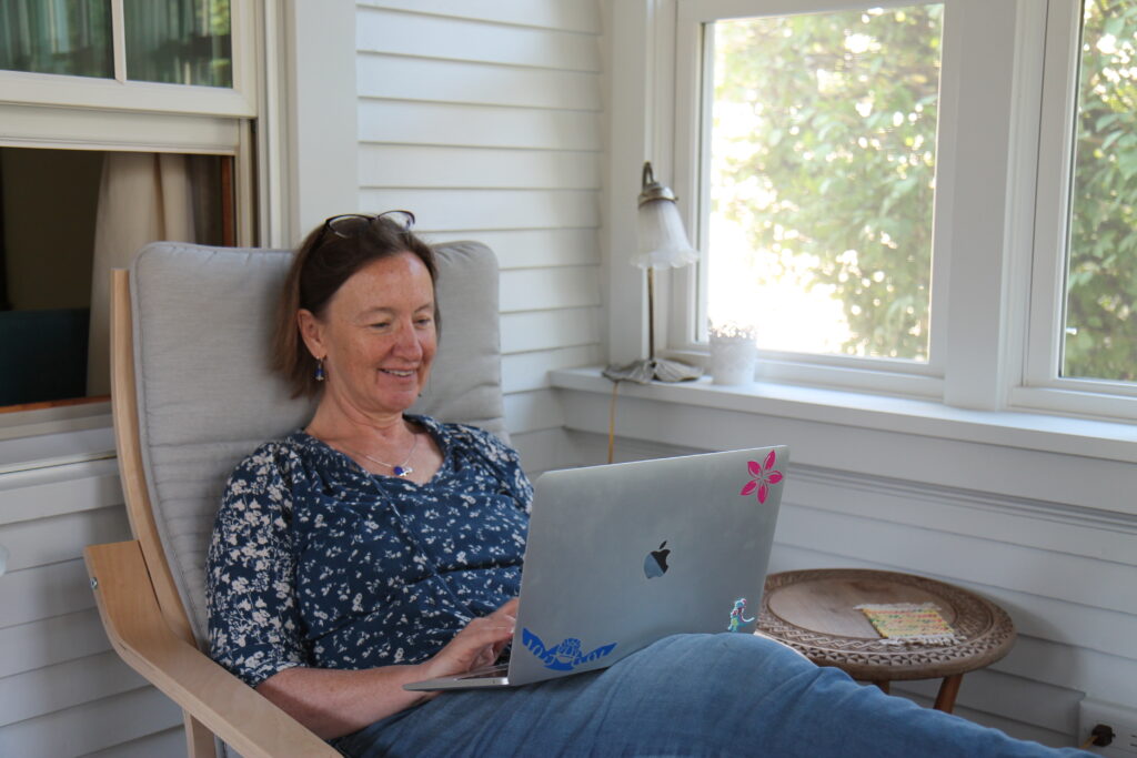 Person wearing blue flowered blouse reclines on porch chair, with silver Apple laptop.