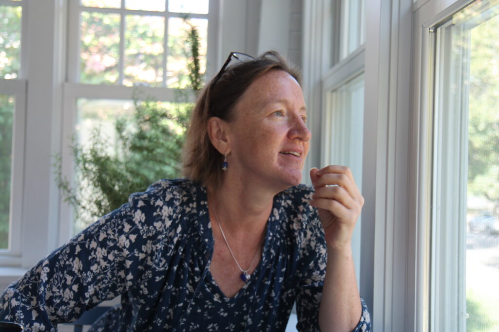 Person with light skin, blue flowered blouse, and glasses on her head looks out porch window and gestures.