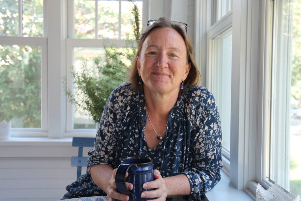 Person with light skin, blue flowered blouse, and glasses holds mug and leans forward, smiling.