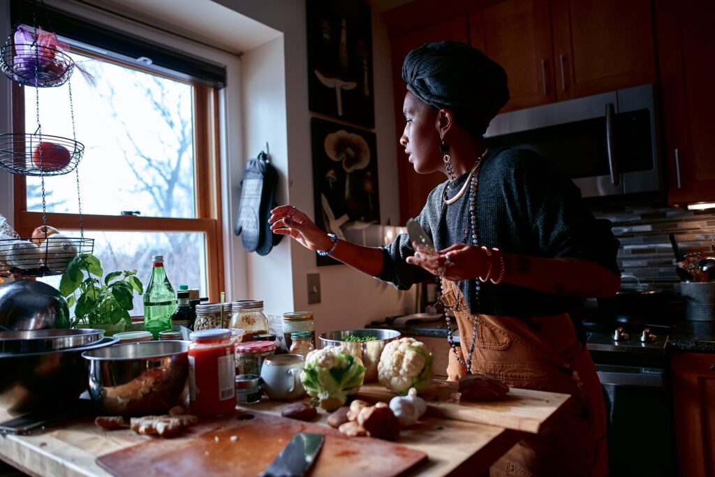 Black woman wearing overalls, jewelry, and gray headwrap reaches across kitchen island full of ingredients.