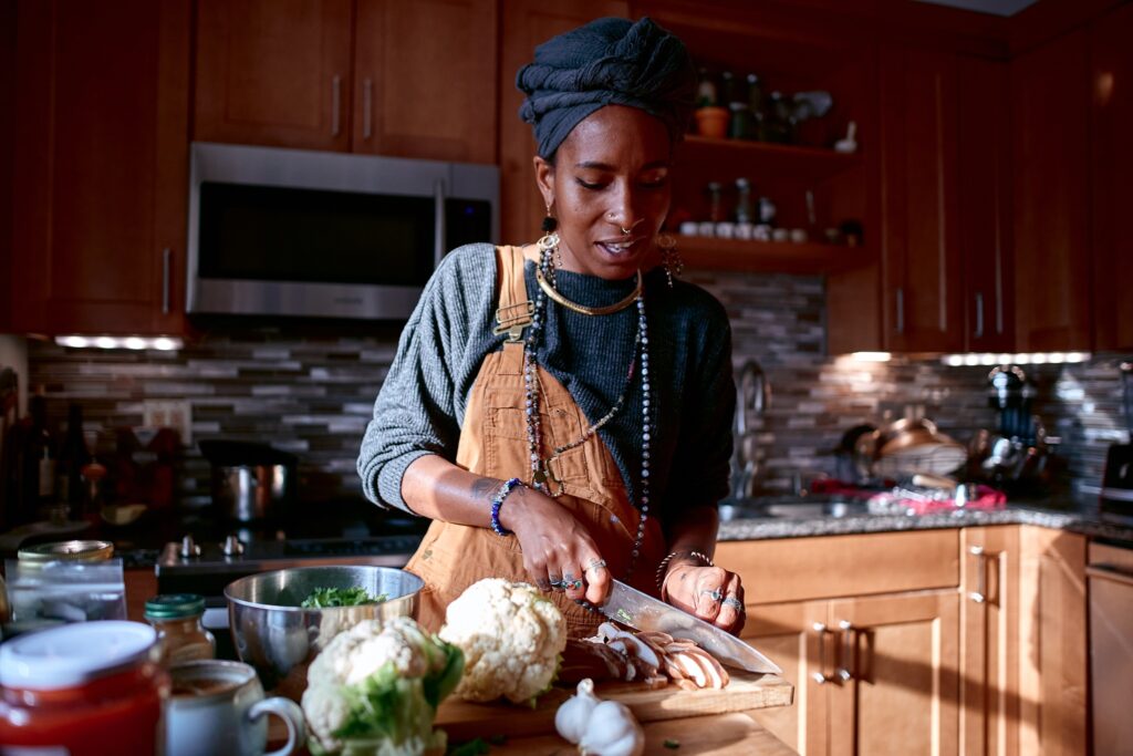 Black woman wearing overalls, jewelry, and gray headwrap smiles slightly while slicing mushrooms.