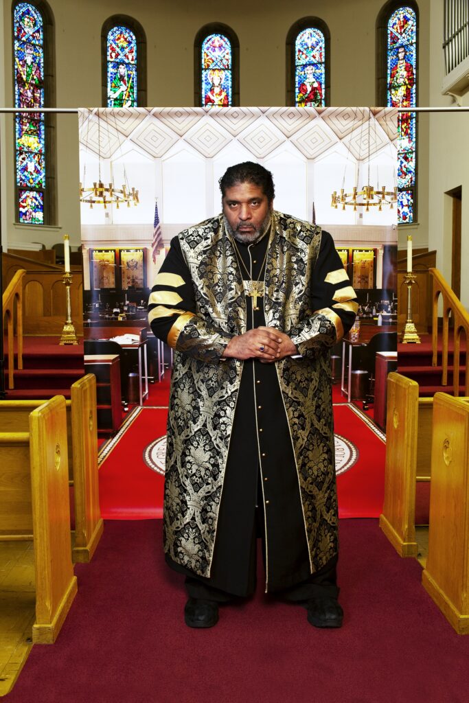 Dark skinned man in gold and black robes stands between church pews with banner behind him.
