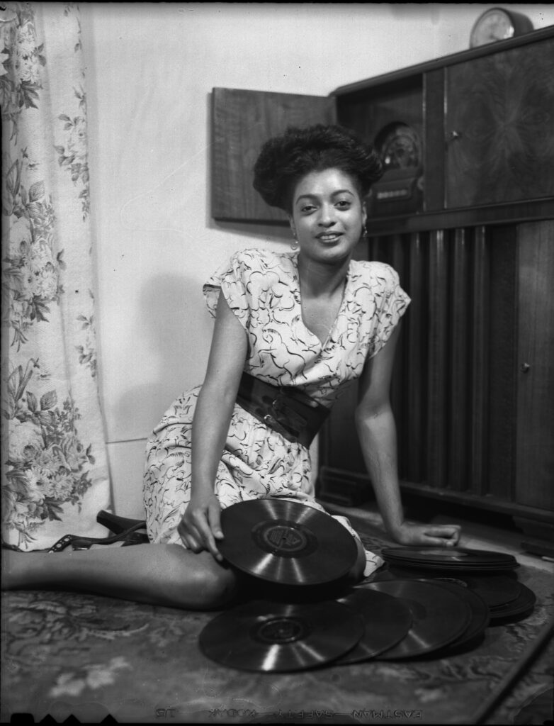 Black and white photograph of a dark skinned woman smiling slightly while sitting on the floor with records around her lap.