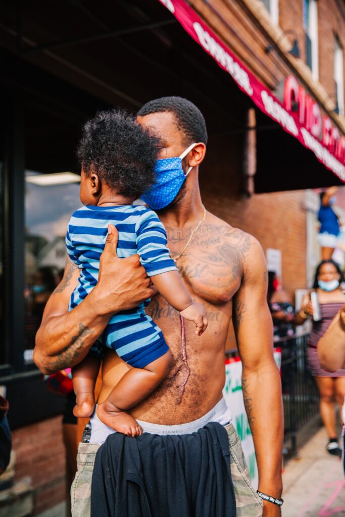 Dark skinned shirtless man wearing a masked looks towards an infant he is holding in one arm.