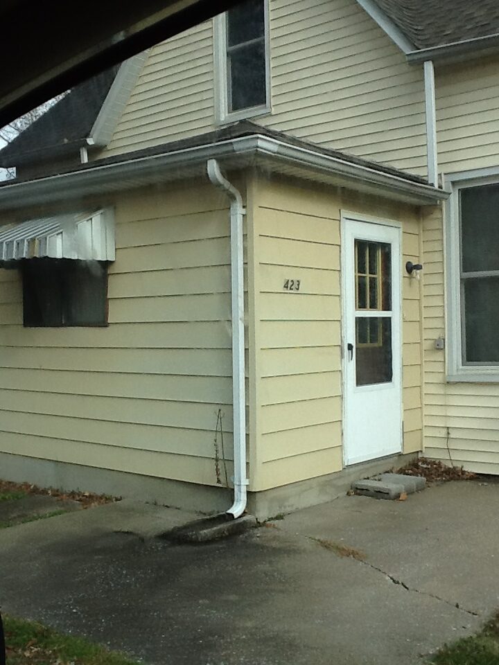 House with yellow siding, white screen door, house number, and awning.