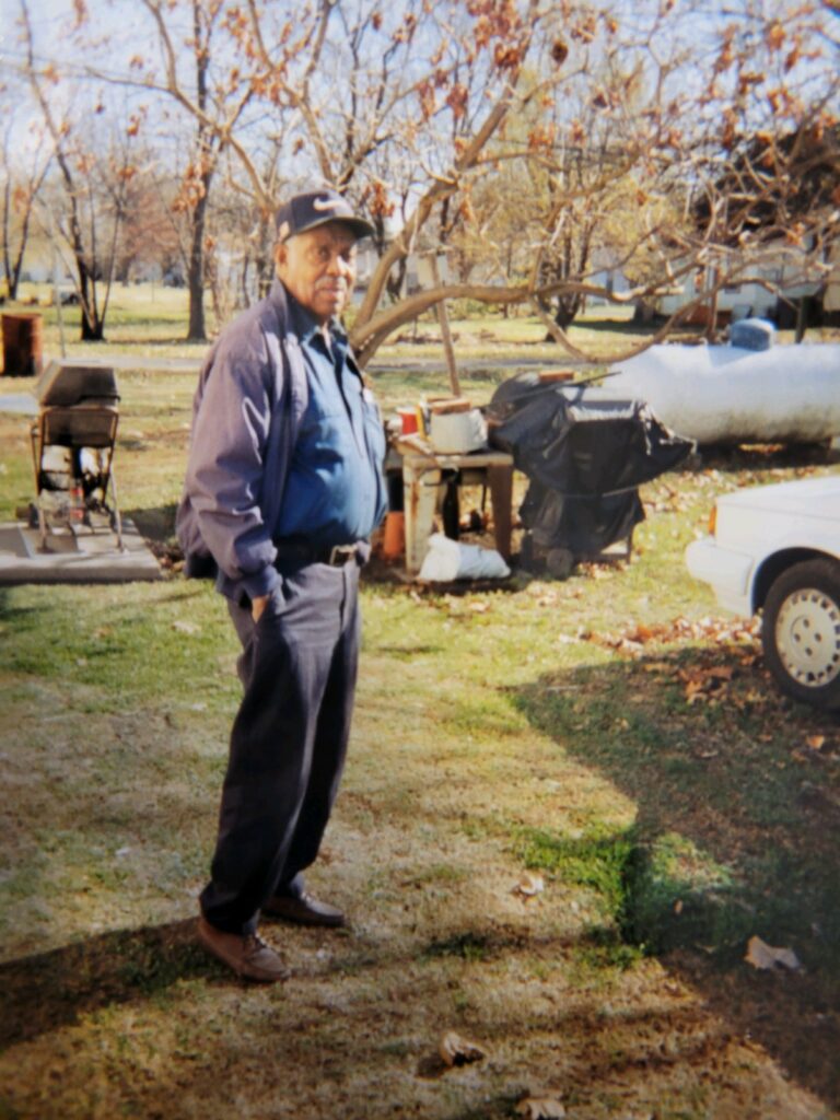 Person with dark skin wearing dark blue and baseball hand stands in cemetery with hands in pockets.