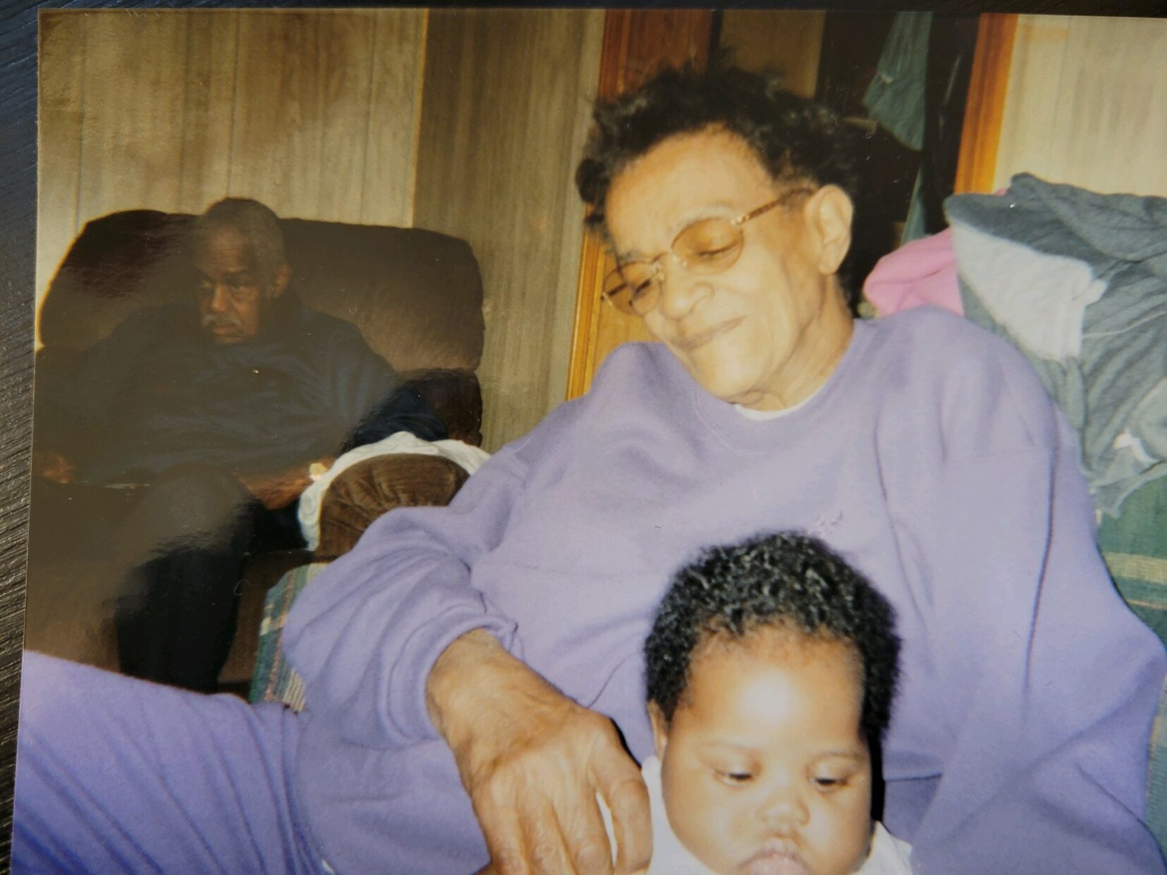 Person with medium dark skin and glasses wears lavender sweats and holds baby in lap, with person in armchair in background.