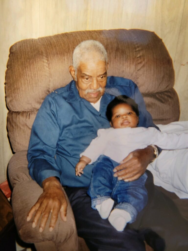 Person with dark skin and gray hair holds infant on lap in brown armchair.