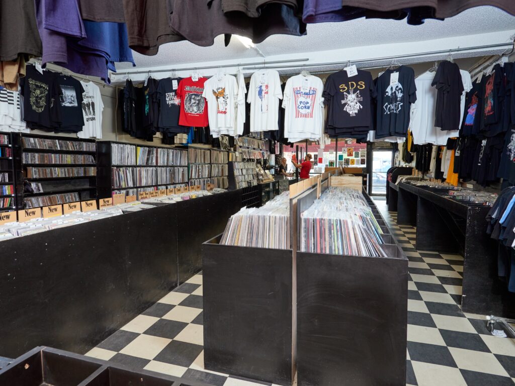 Interior of record store with bins of records, hanging t-shirts, and black and white tiled floor.