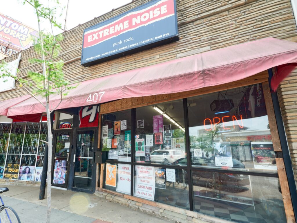 Storefront with red awning, many posters in glass windows, and black and red sign reading EXTREME NOISE.