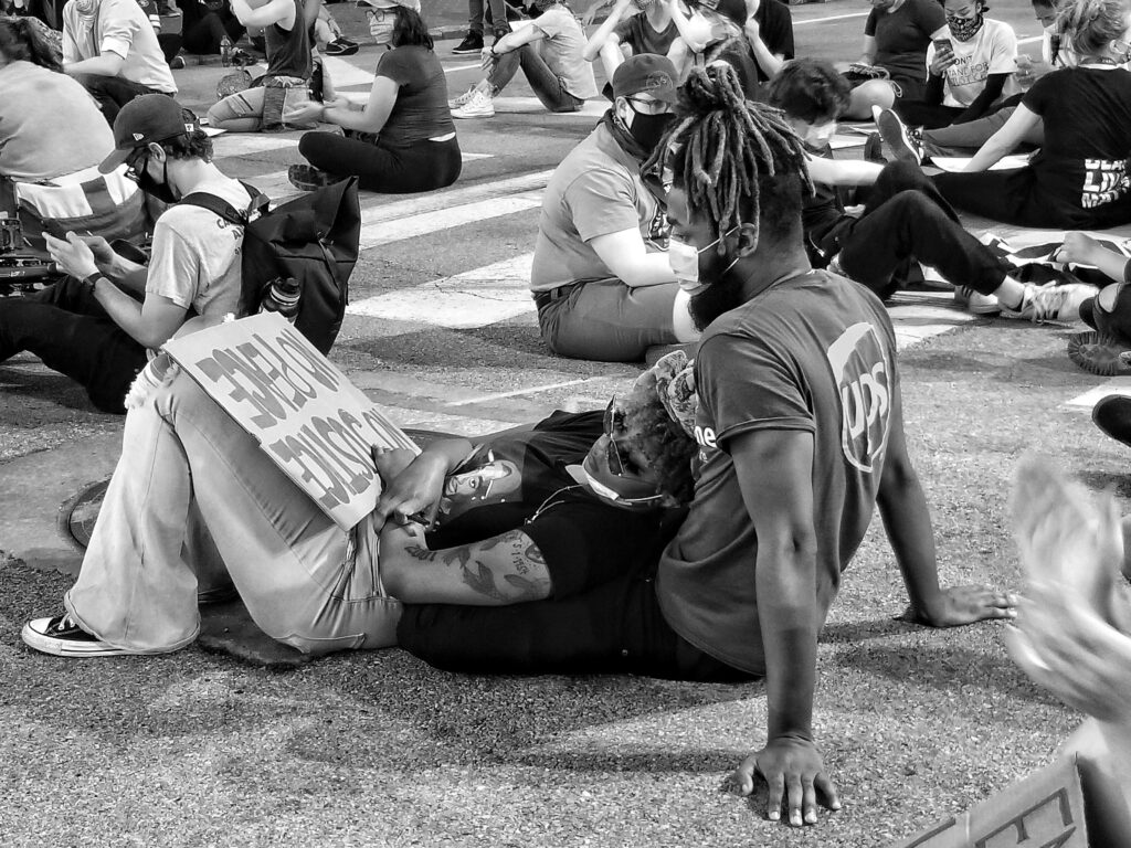 Black and white image of a masked couple leaning against each other with a protest sign, with many people sitting on pavement around them.