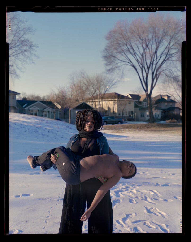 Dark skinned woman holds a young boy in her arms in winter with houses in the background