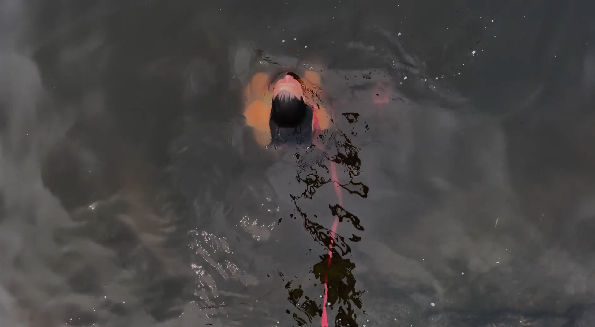 Aerial view of naked person with bright pink ribbon submerged in dark water with head emerging