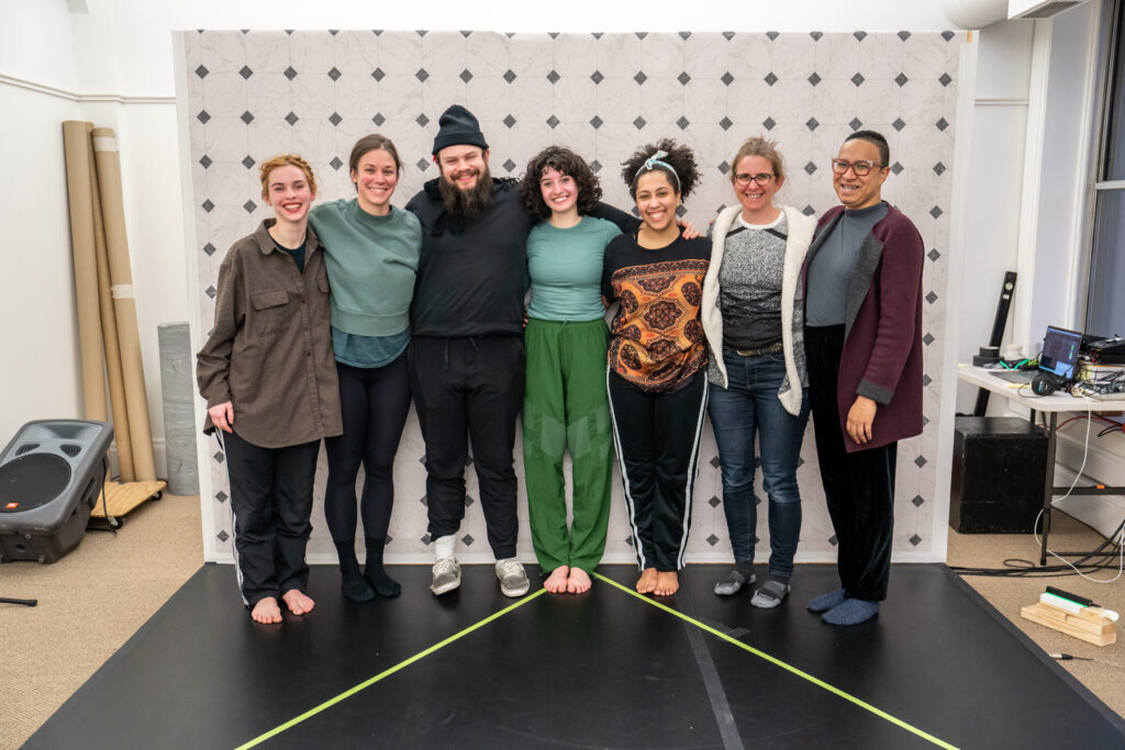 Seven people stand smiling with arms around each other, on set with wall and dance floor.