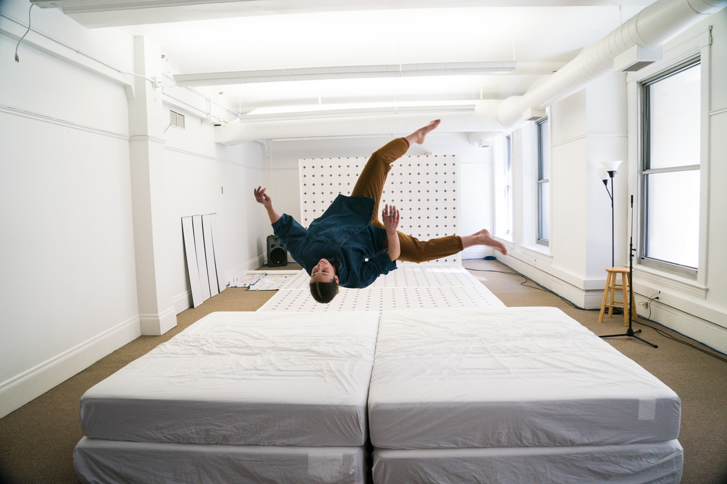 Person in blue shirt and brown pants falling backward in mid air over two white mattresses.