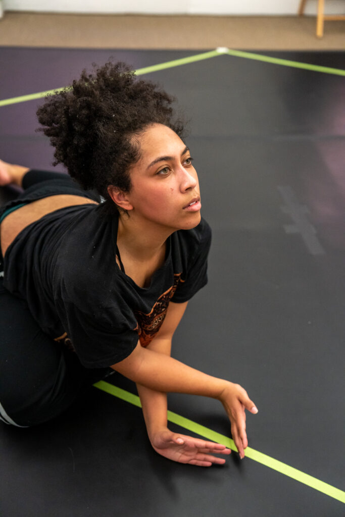 Person with medium dark skin and curly hair in ponytail moves on her belly on marley floor, with hands crossed and eyes looking up.