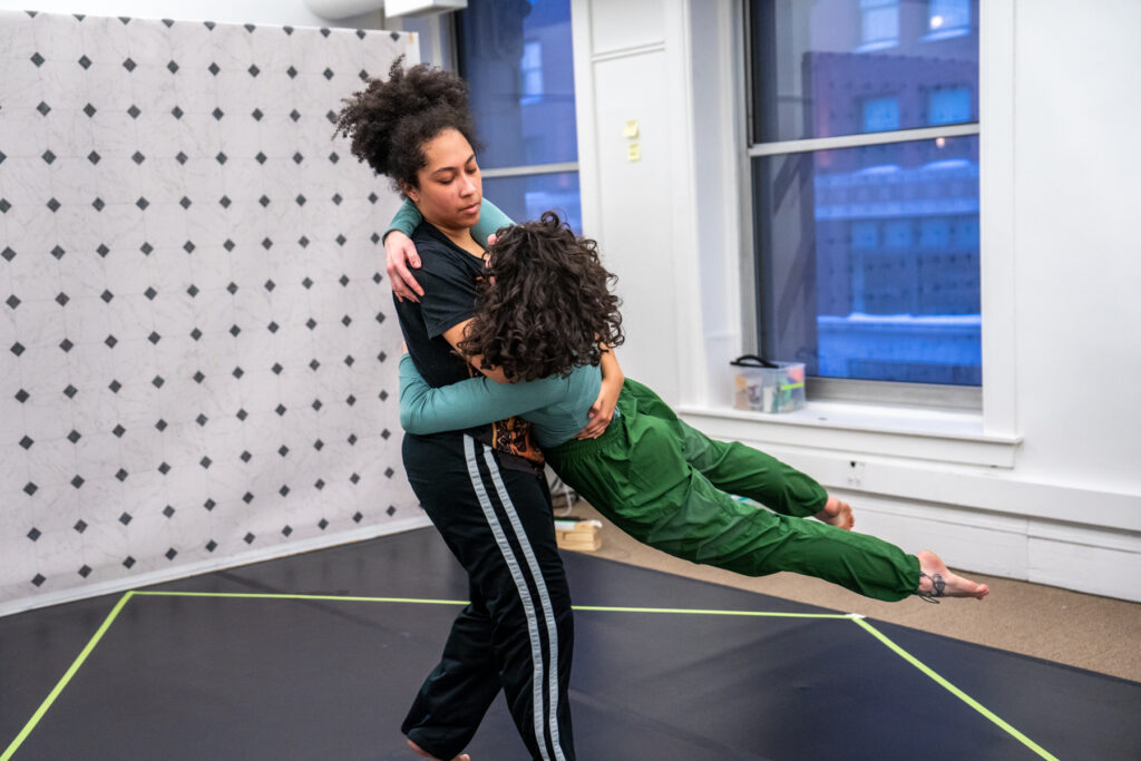 Two dancers with curly hair wrap their arms around each other, one swinging the other's feet off the ground.
