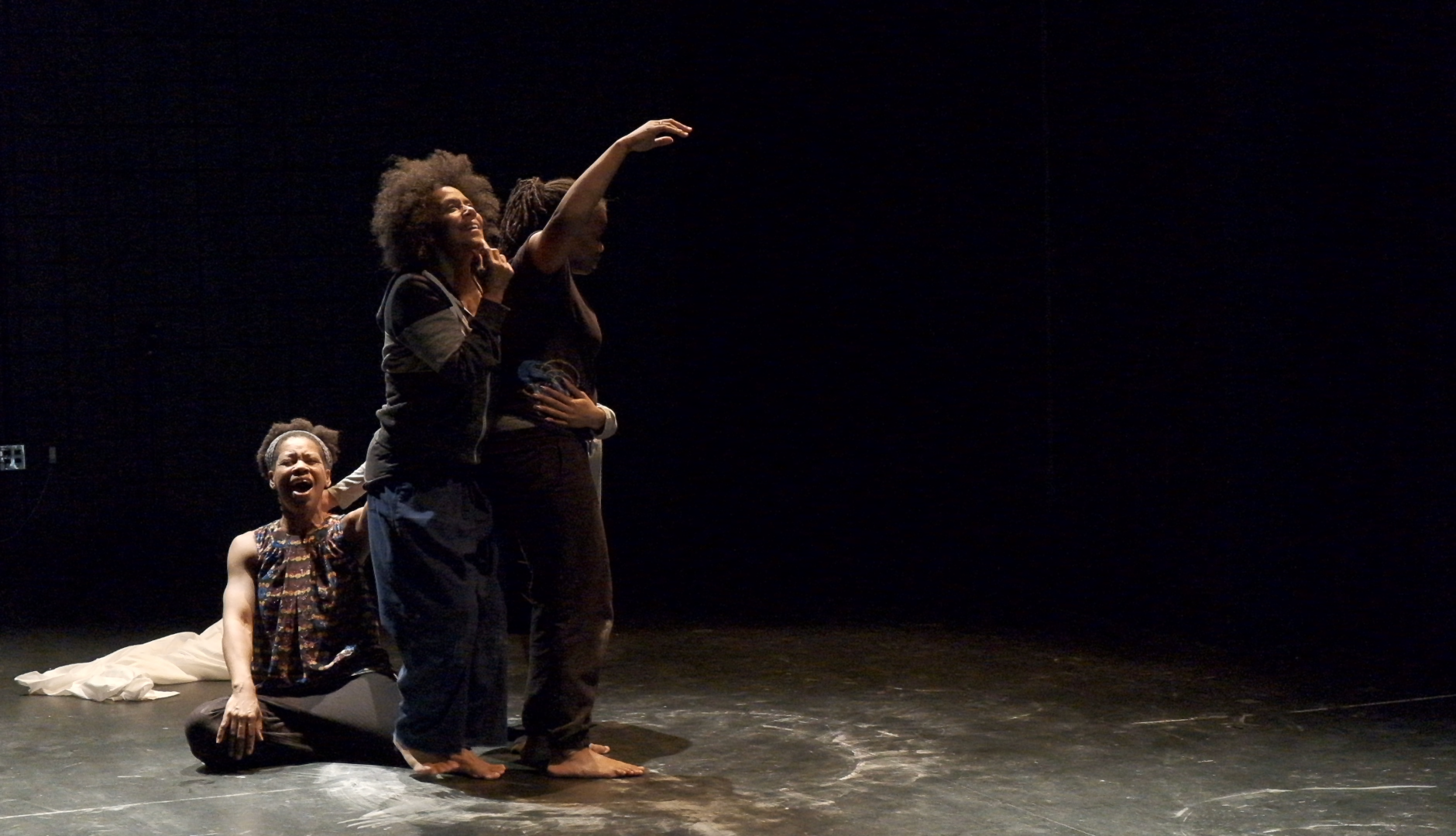 Four people with dark skin are lit on dark stage. One is sitting with mouth open, one is reaching, one is looking up, and another is embracing the others.