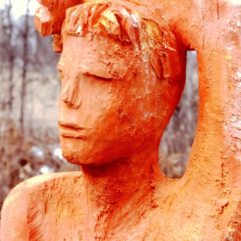 Carved wooden sculpture of human form painted orange, with arm resting on top of head.
