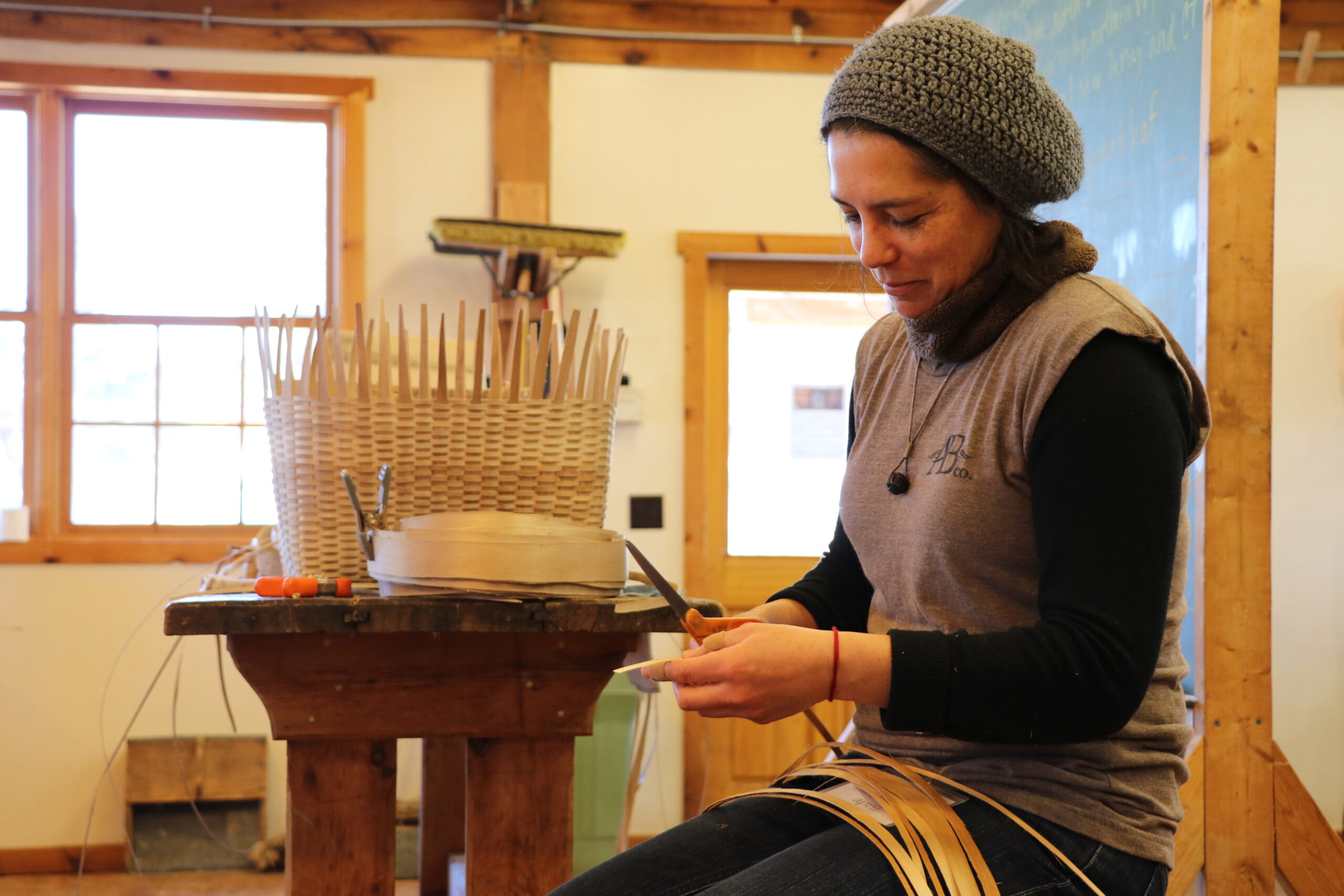 Person wearing knit hat and scarf cuts strip of wood with scissors, with in-progress basket on table in front of them.