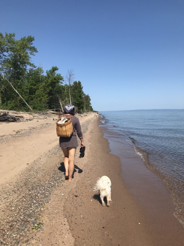 Person walks down lakeshore wearing basket backpack, white white dog beside them.