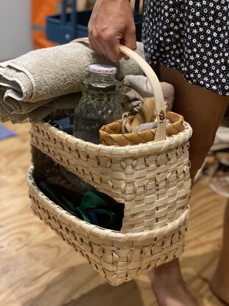 Person holds woven basket, filled with glass bottle, towel, and other items.