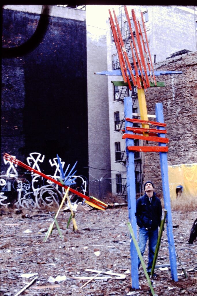 Person looks up at large wooden sculptures with animal-like forms in primary colors, in empty city lot.