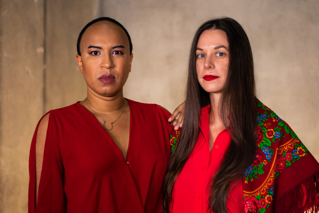 Two people wearing red lipstick and red shirts stand close and look directly at the camera, one with medium dark skin and short hair and the other with light skin and long hair.