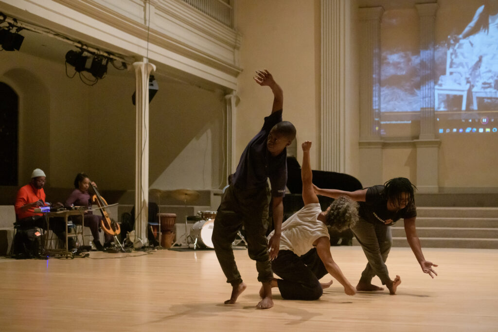 Three dancers with dark skin make various shapes in proximity to each other, with two musicians and projected image behind them.